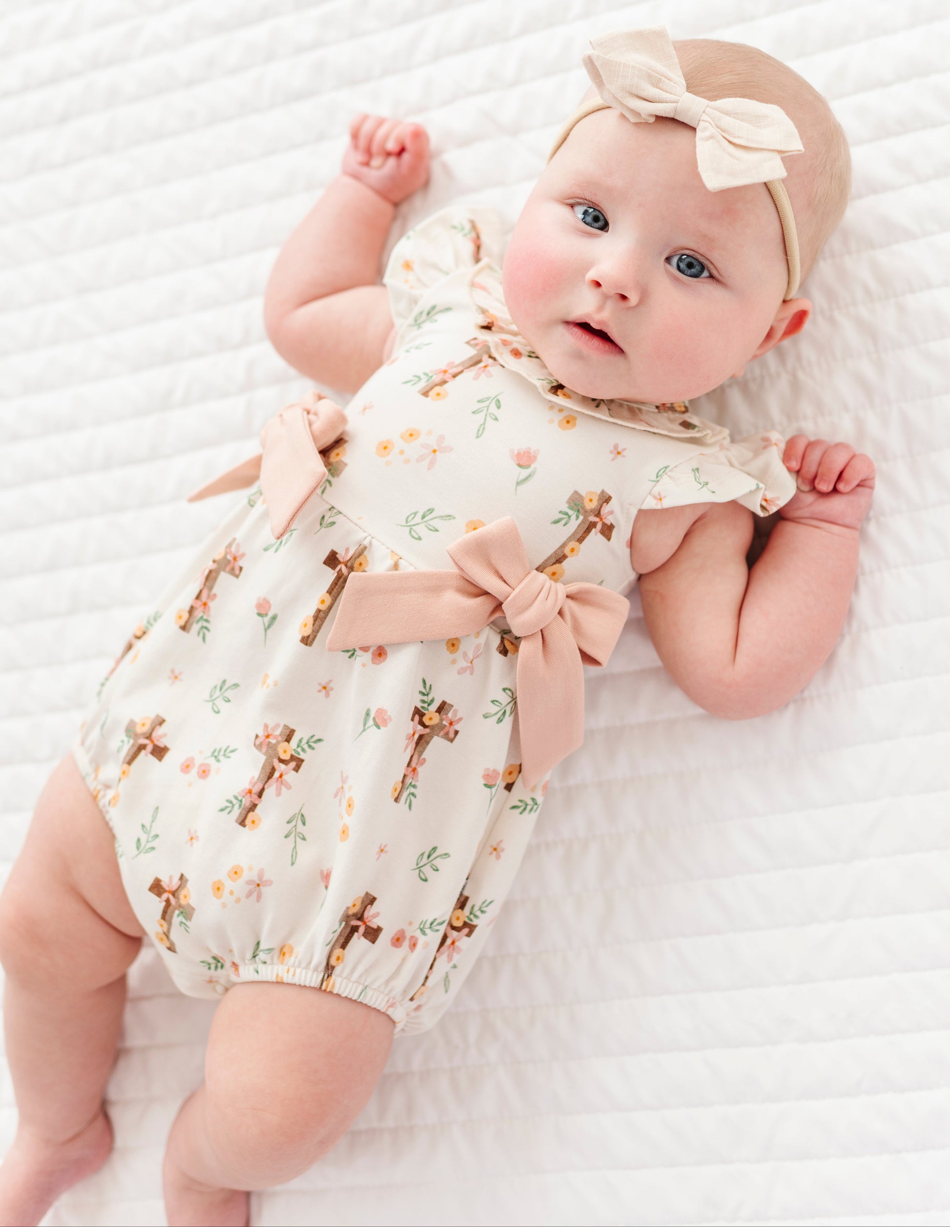 baby girl in a cross with florals bubble romper and a neutral colored headband on a white background 