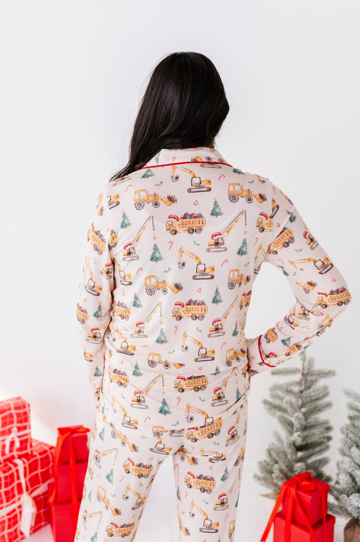 A woman viewed from behind wearing a matching long-sleeve pajama set with an all-over Christmas construction theme print, featuring yellow dump trucks, cranes, and snowmobiles on a cream background. She is standing in a holiday setting with wrapped gifts and a small Christmas tree.
