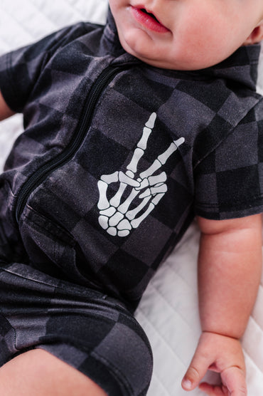A close-up of a baby wearing a black and dark gray checkered zip-up romper with a white skeleton hand graphic making a peace sign, lying on a white quilted surface.