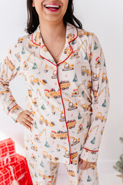A woman wearing a long-sleeve bamboo pajama set with a holiday construction theme, featuring yellow excavators, dump trucks, and Christmas trees on a cream background with red piping.