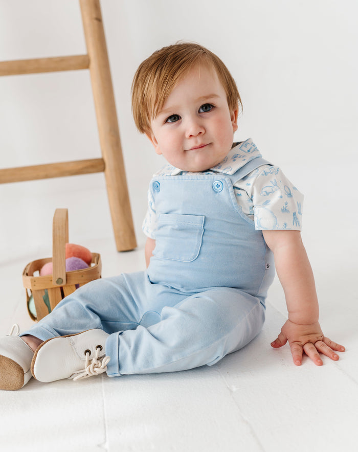 Baby wearing a light blue outfit sitting on a white floor with a wooden ladder in the background.