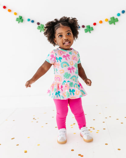 Young girl wearing a lucky charm bamboo cotton peplum top with a pattern of rainbows and bows, standing on a white background with confetti.