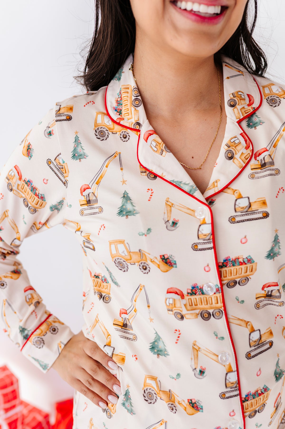 A close-up of a woman wearing long-sleeve bamboo pajama top with an all-over construction vehicle holiday print on a cream background with red piping.