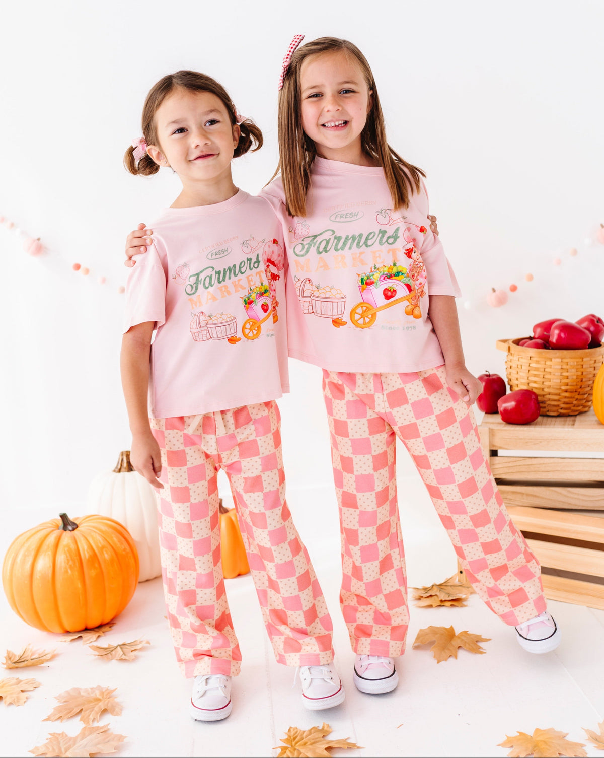 Two young girls wearing matching pink short-sleeve t-shirts and pink-and-white checkered pajama pants, posed in a fall-themed studio setting with pumpkins, leaves, and baskets of apples.