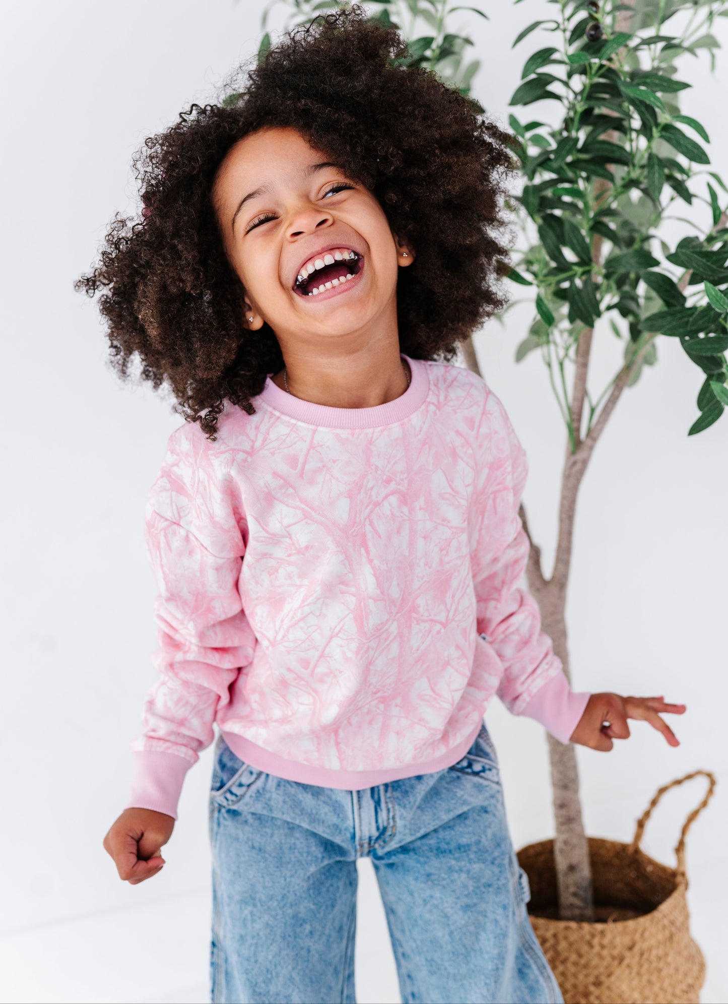 Child wearing a pink sweatshirt with pink camo patterns, standing in front of a plant.