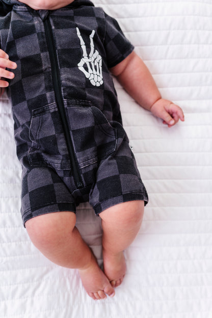 A baby lying on a white quilted blanket wearing a black and gray checkered zip-up romper with a white skeleton hand graphic on the chest.