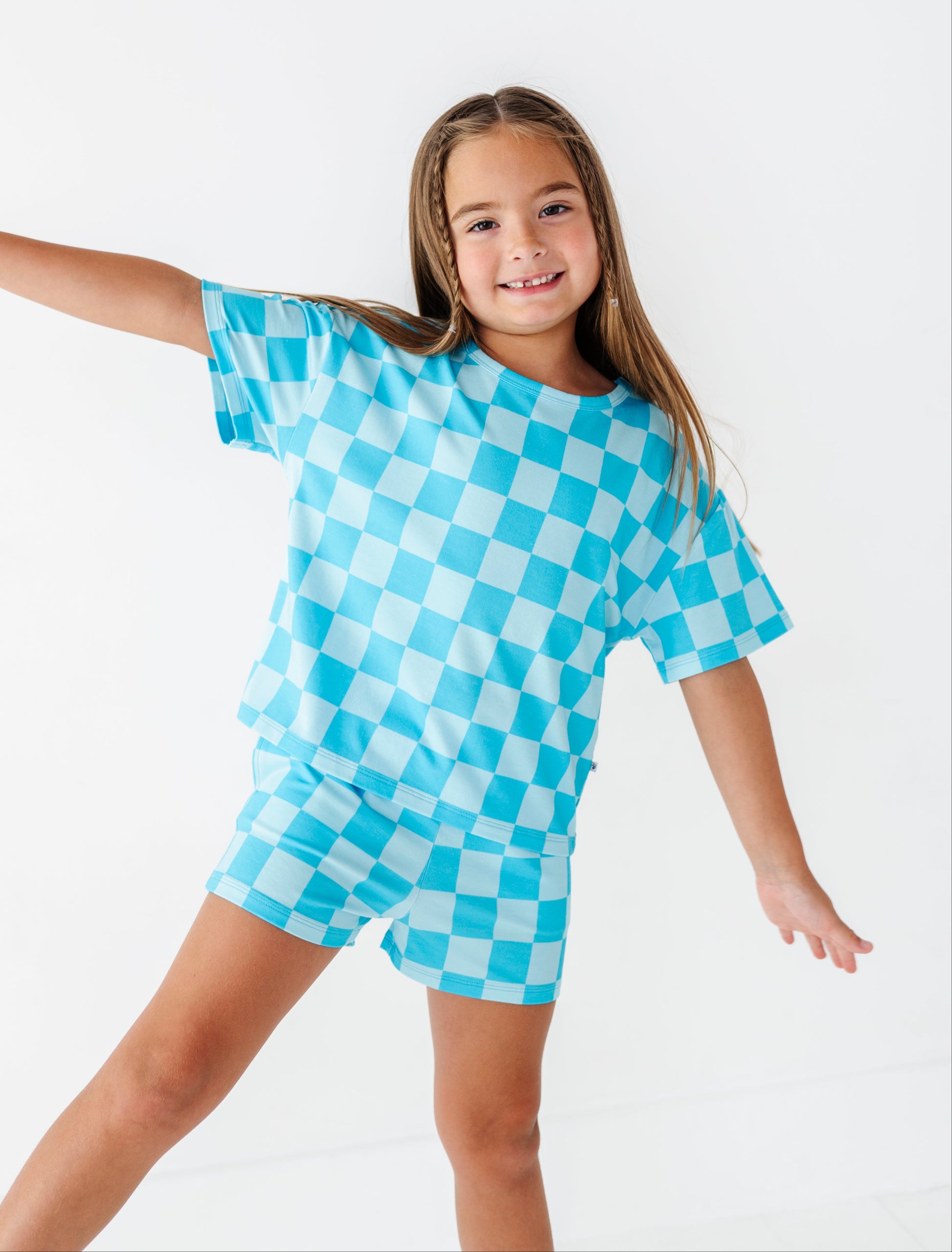 A young girl with long light brown hair is modeling a matching two-piece lounge set featuring a bright turquoise and white checkerboard pattern. She is posing against a seamless white studio background.