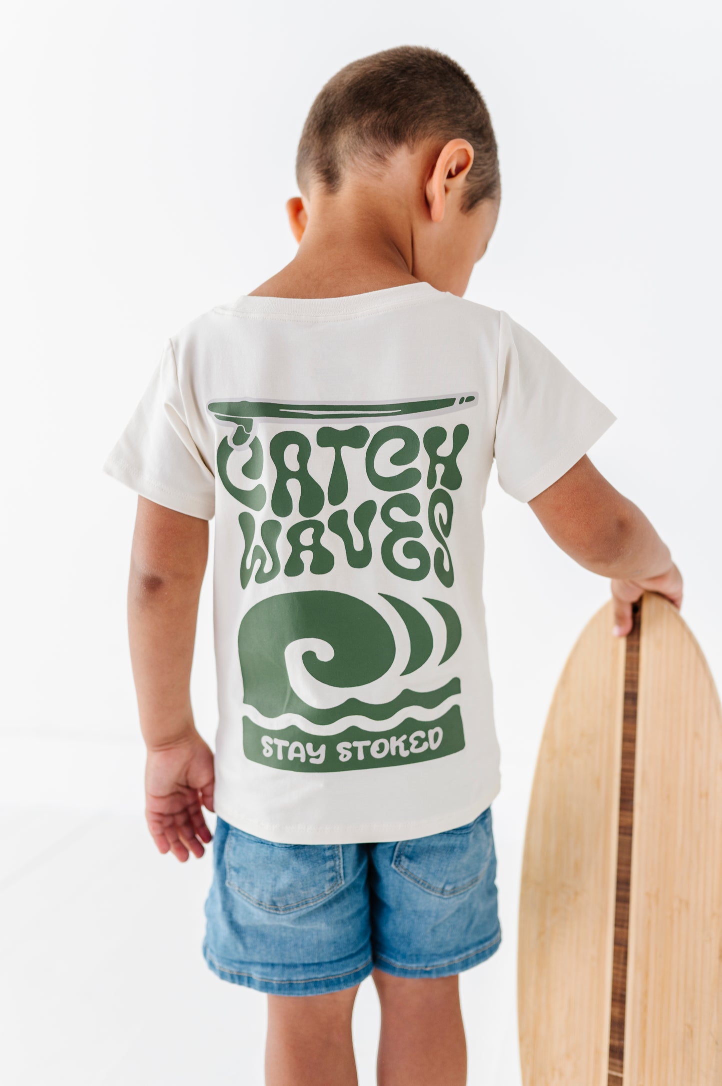 Child wearing a white t-shirt with green text and graphics, standing next to a wooden surfboard.