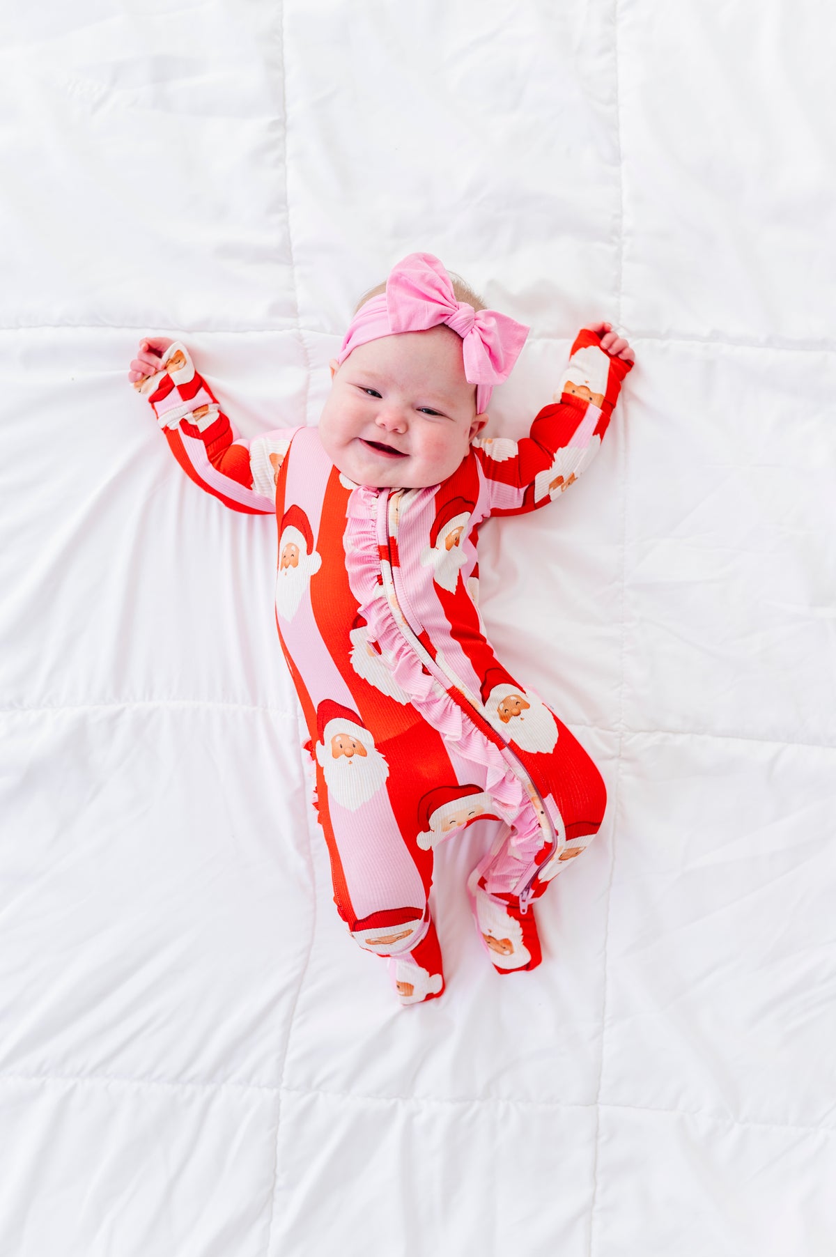 A baby lying on a white quilted blanket, wearing a red and white Santa Claus print footed bamboo sleeper with a pink bow headband.