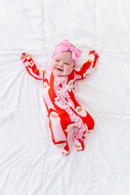 A baby lying on a white quilted blanket, wearing a red and white Santa Claus print footed bamboo sleeper with a pink bow headband.