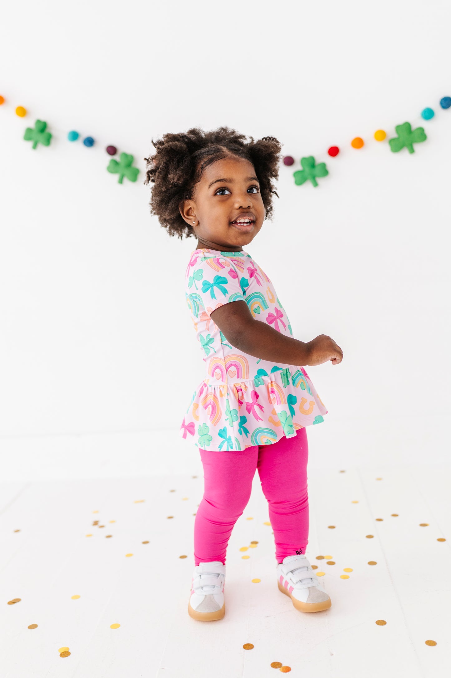 Child wearing a St. Patrick's Day bamboo cotton outfit with pink leggings, standing on a white background with confetti.