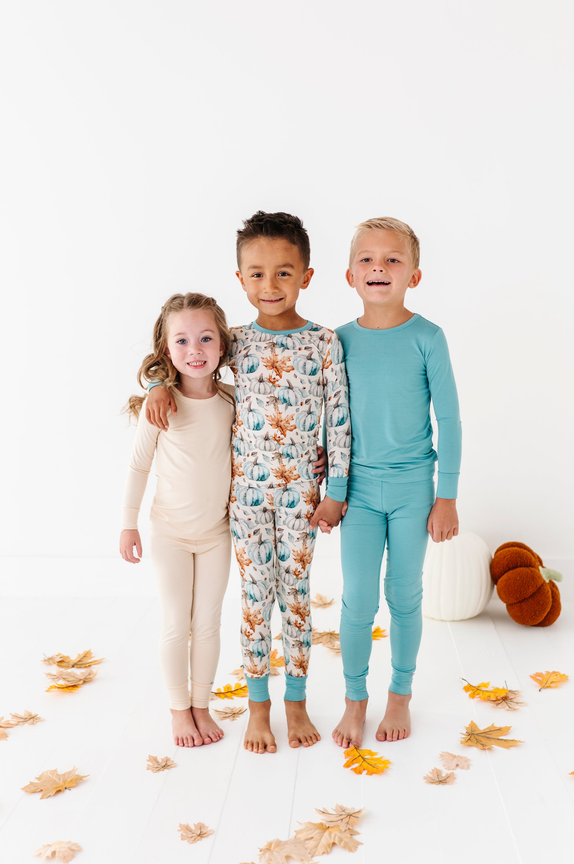 Three children wearing bamboo ajamas standing together on a white background with autumn leaves.