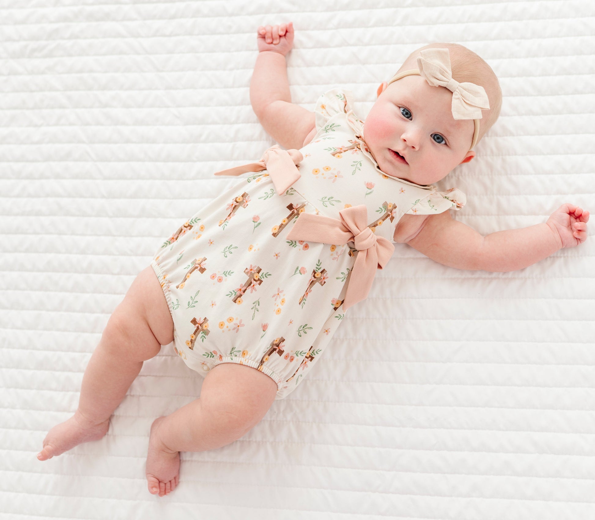 baby girl in a cross with florals bubble romper and a neutral colored headband on a white background 
