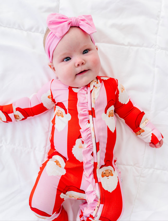 A baby lying on a white quilted blanket, wearing a red and pink Santa Claus print footed bamboo sleeper with a pink ruffle zipper and a matching pink bow headband.