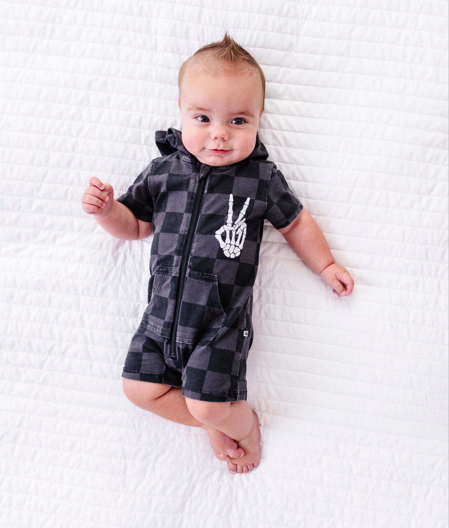 A baby lying on a white quilted blanket, wearing a black and gray checkered short-sleeve romper with a white skeleton hand graphic on the chest.