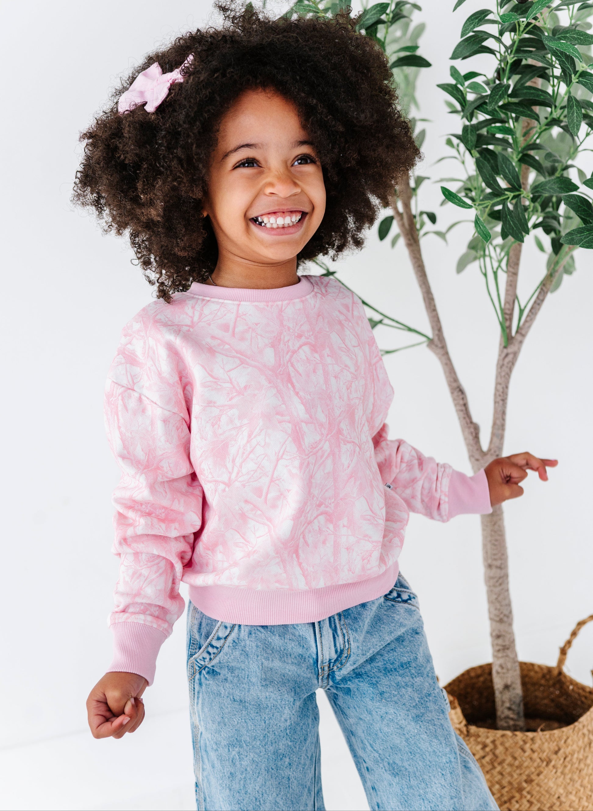 Child wearing a pink tree camo sweatshirt and blue jeans, standing next to a plant.