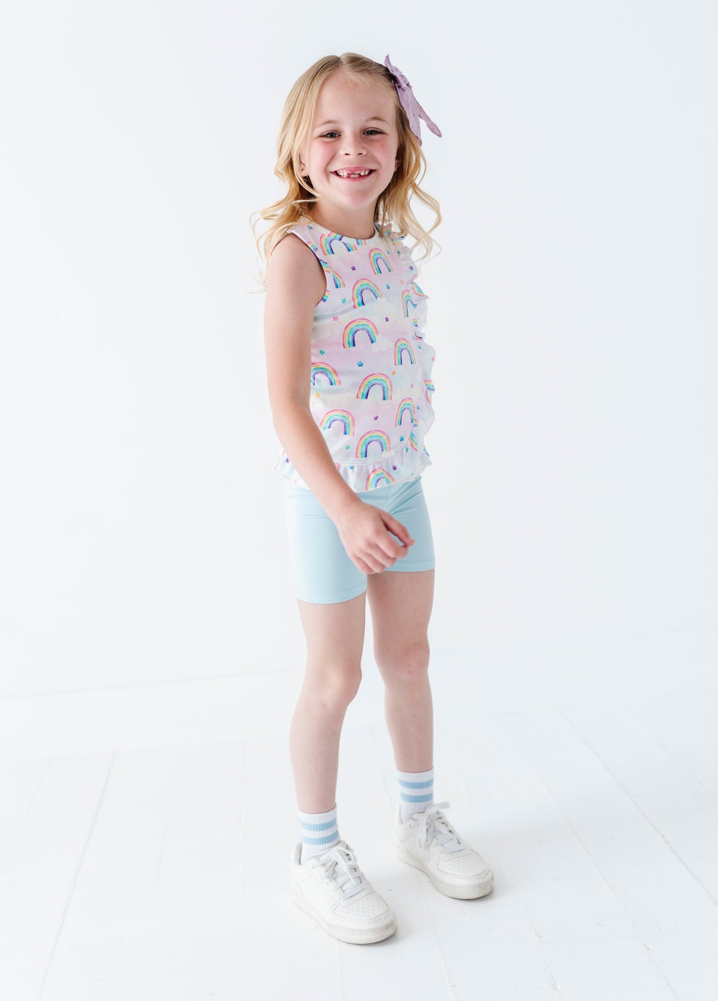 Young girl wearing a sleeveless bamboo cotton peplum top with watercolor rainbow pattern and light blue bamboo biker shorts on a white background
