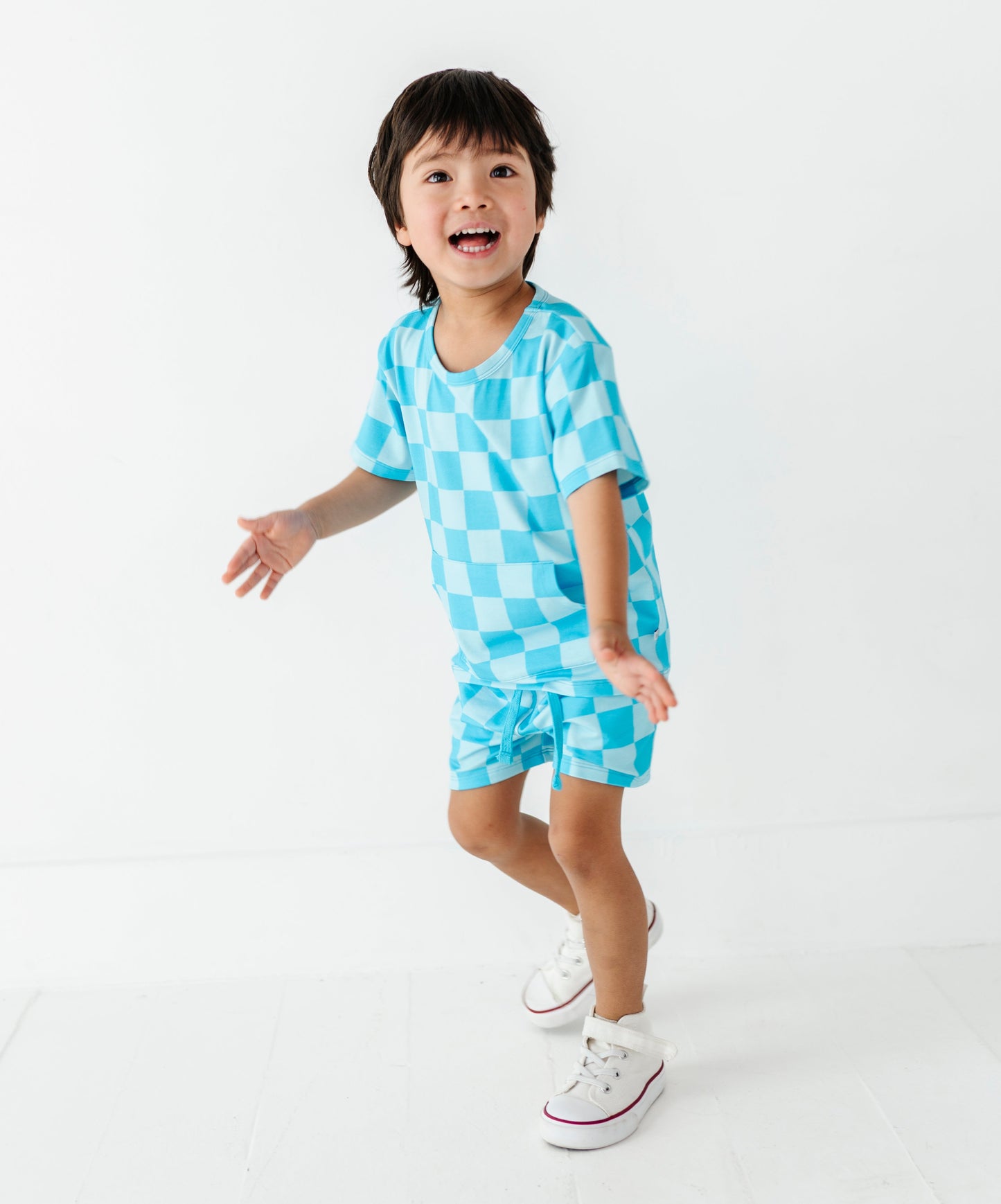 A young boy wearing a light blue checkered short-sleeve t-shirt and matching shorts, posing in a playful stance against a clean white studio background.