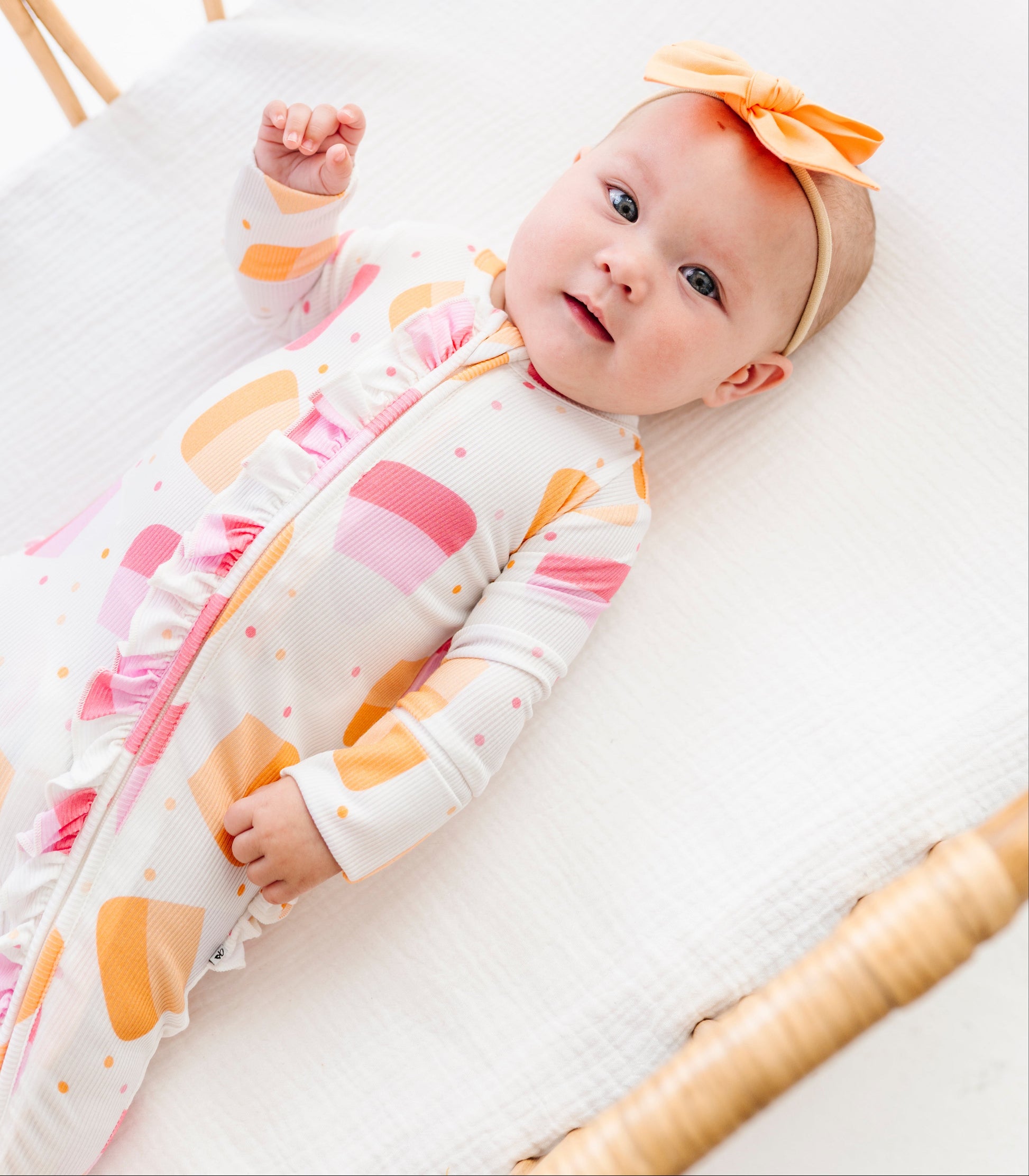 A baby lying on a white muslin mattress in a rattan bassinet, wearing a white footless sleeper with pink and orange abstract shapes and a matching orange bow headband.