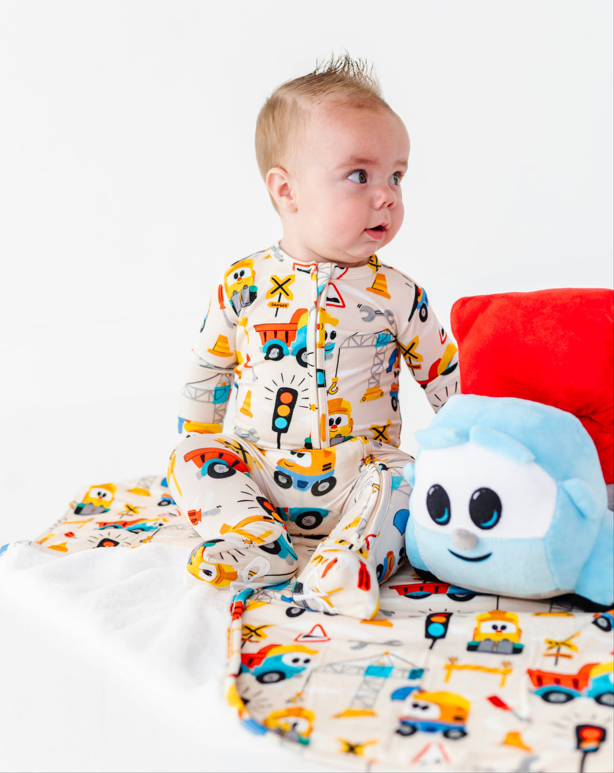 A baby sitting on a white blanket wearing a cream-colored footed pajama suit with a colorful construction vehicle print. The baby is posed next to a blue plush toy and a red pillow on a clean white background.