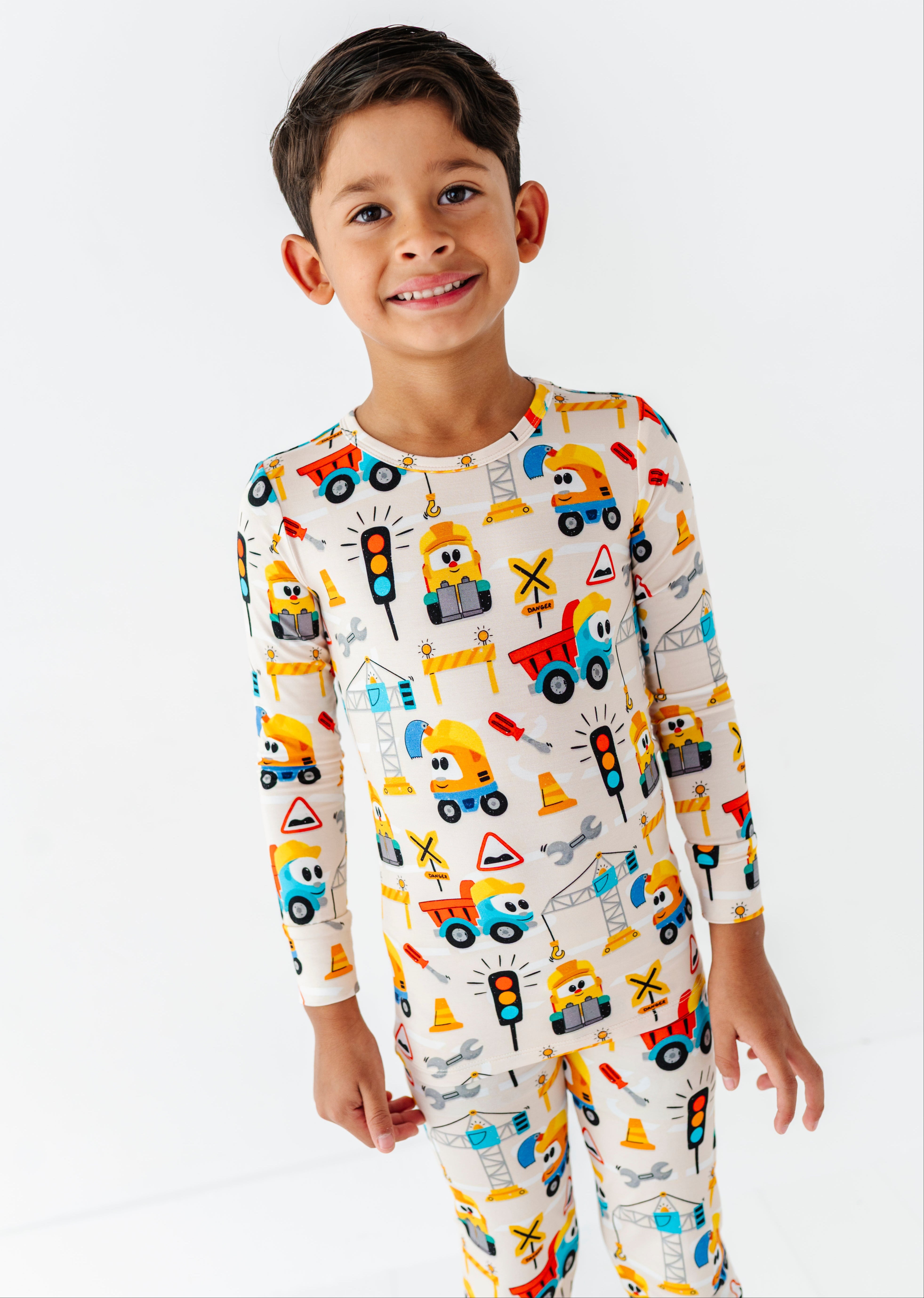 A young boy smiling and wearing a long-sleeve pajama set with a colorful construction vehicle print on a white background.
