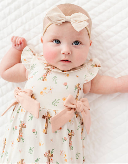 baby girl in a cross with florals bubble romper and a neutral colored headband on a white background 