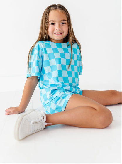 A young girl with braided hair sits on a white floor wearing a light blue and white checkerboard short-sleeve dress and white sneakers against a white studio background.