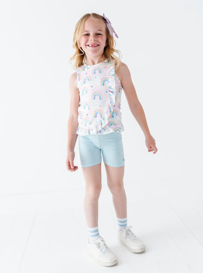 Young girl wearing a sleeveless bamboo cotton peplum top with watercolor rainbow pattern and light blue bamboo biker shorts on a white background