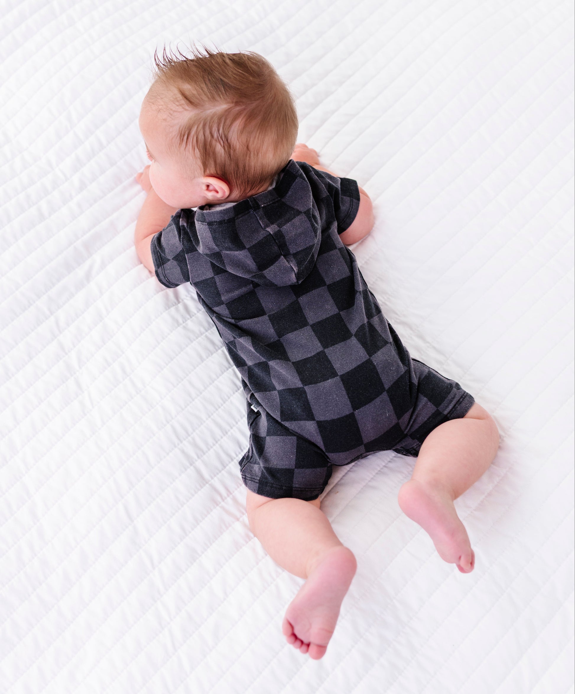 A baby lying on their stomach on a white quilted blanket, wearing a short-sleeve hooded romper in a dark gray and black checkerboard pattern.