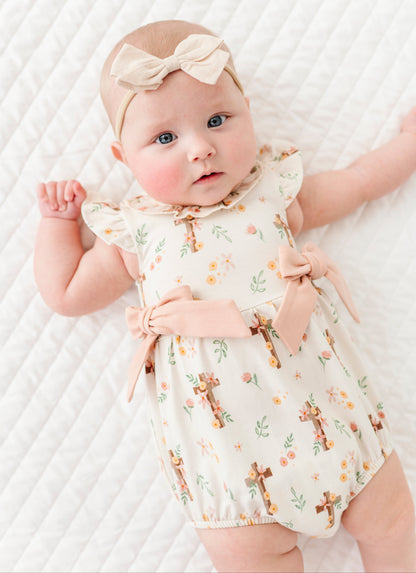 baby girl in a cross with florals bubble romper and a neutral colored headband on a white background 