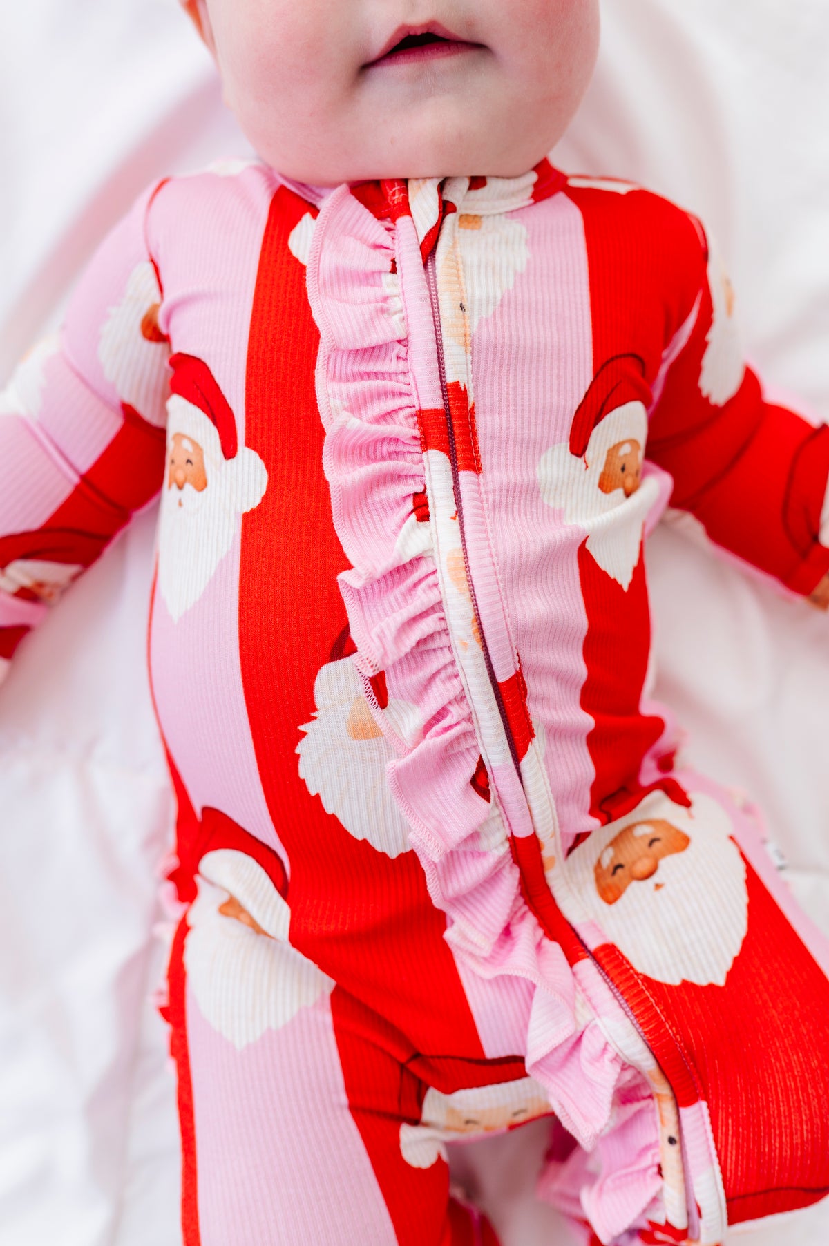 A close-up of a baby wearing a red and pink striped ribbed one-piece bamboo sleeper with Santa Claus face prints and a ruffled front zipper.