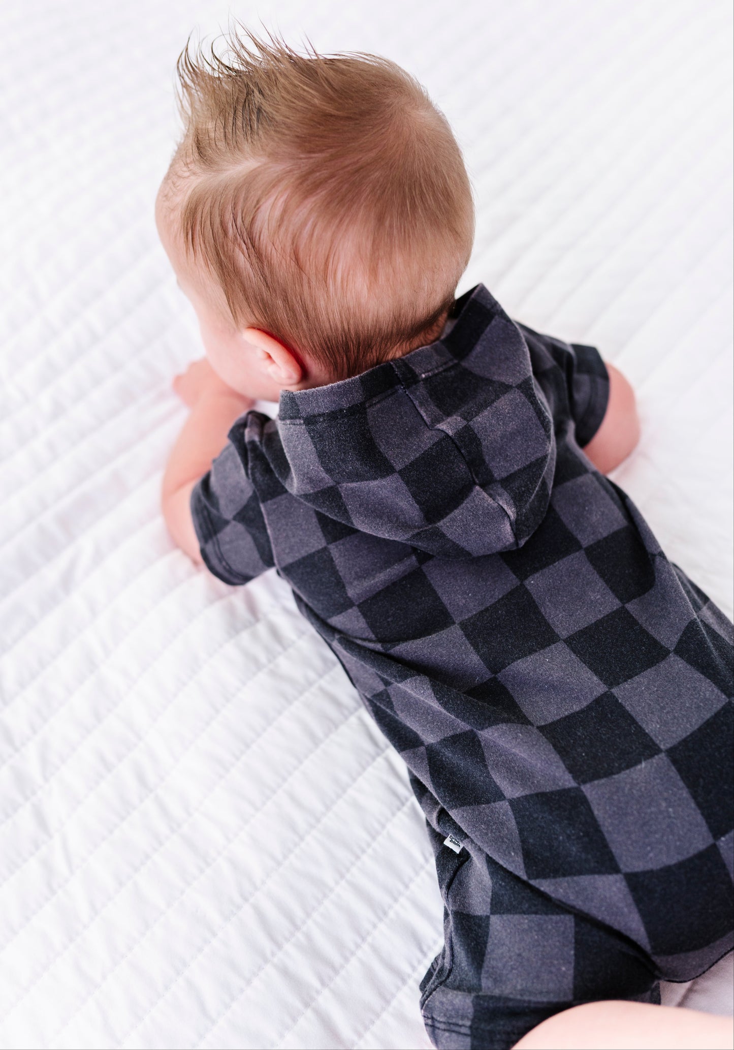 A baby lying on a white quilted blanket, viewed from behind, wearing a dark gray and black checkered hooded short-sleeve romper.