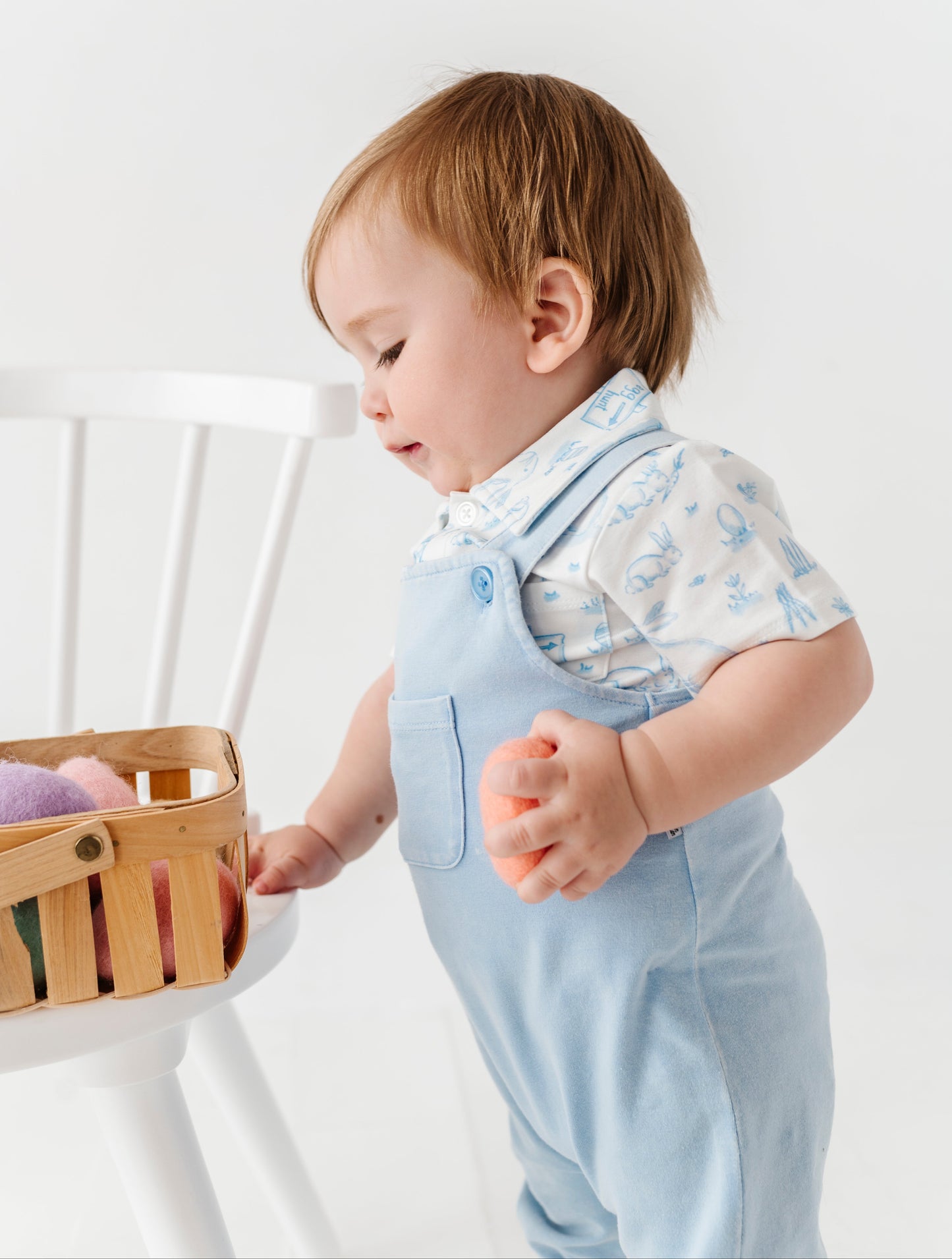 Child wearing light blue overalls and a patterned shirt, standing next to a white crib with a basket.