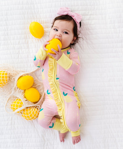 Baby in a pink and yellow romper holding a lemon-shaped toy on a white textured surface