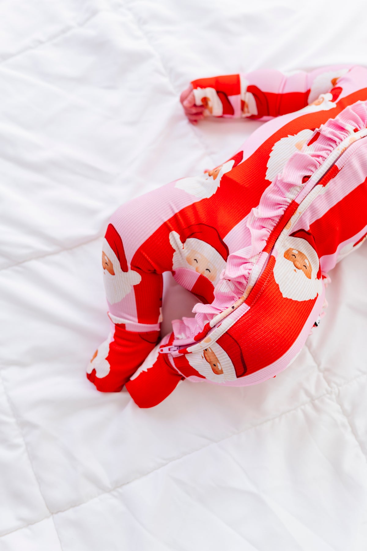 A baby's footed bamboo sleeper in red and pink with a Santa Claus print, featuring ruffled trim, laid out on a white quilted blanket.