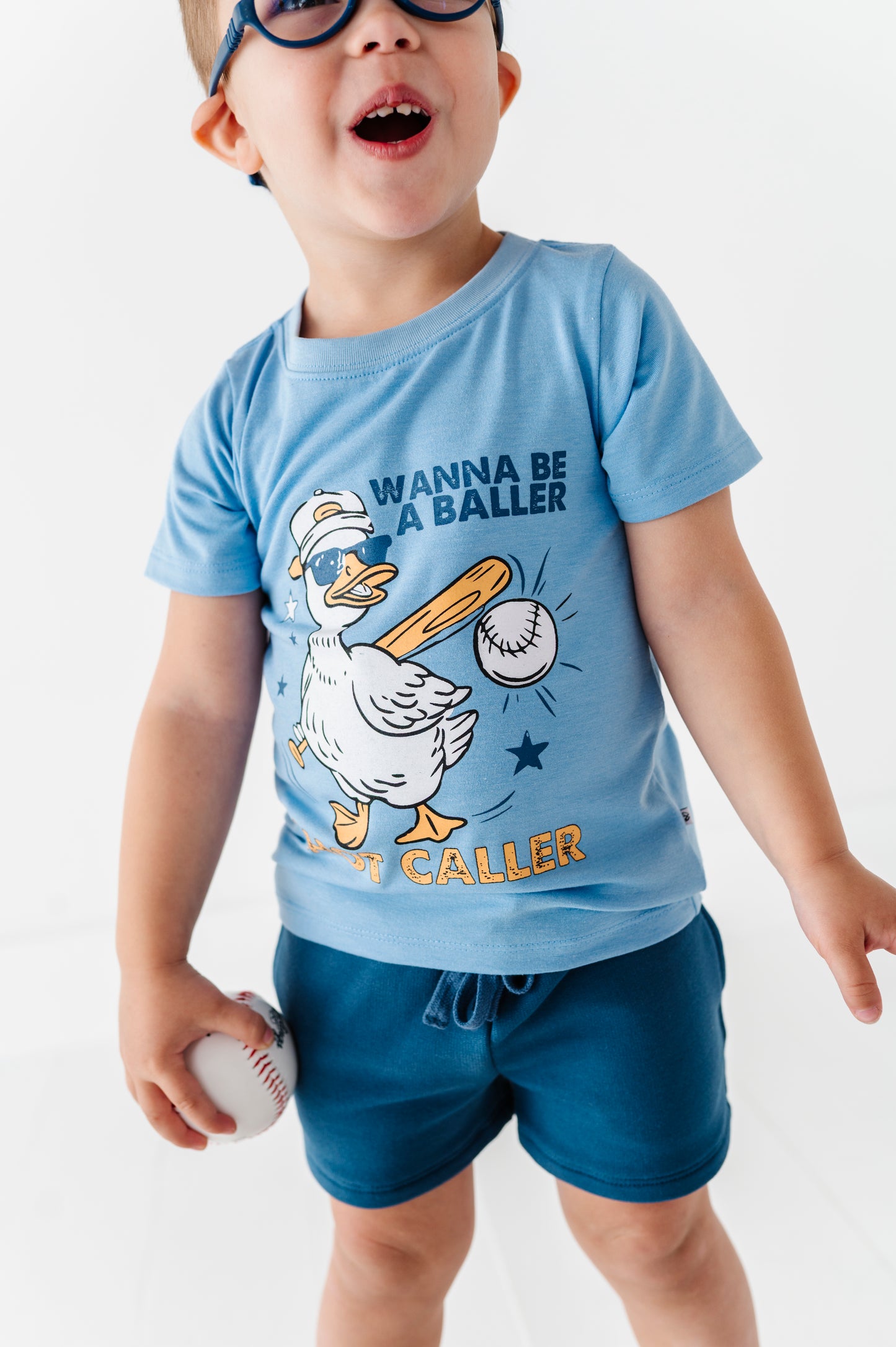 Child wearing a baseball-themed blue t-shirt with a cartoon duck graphic and text, standing against a white background.