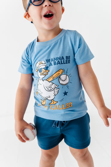 Child wearing a baseball-themed blue t-shirt with a cartoon duck graphic and text, standing against a white background.