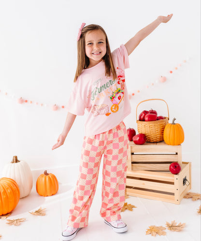 A young girl modeling a light pink short-sleeve t-shirt and matching pink-and-white checkered pajama pants in a fall-themed studio setting with pumpkins, apples, and wooden crates.