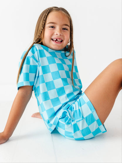 A young girl with braided hair sits smiling on a white floor, wearing a short-sleeve dress with a light blue and white checkerboard pattern.
