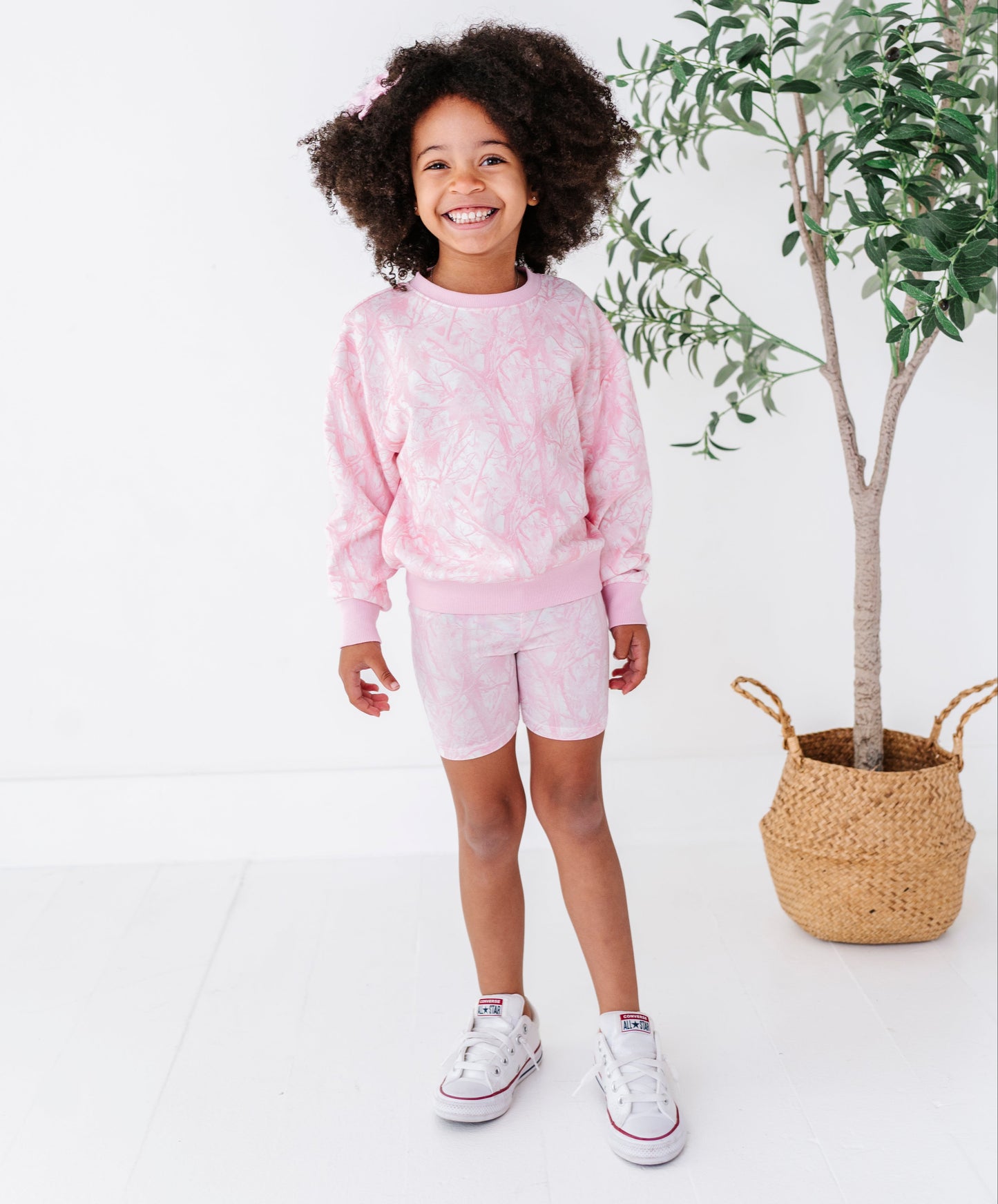Child wearing a pink tree camo outfit standing next to a plant on a white background
