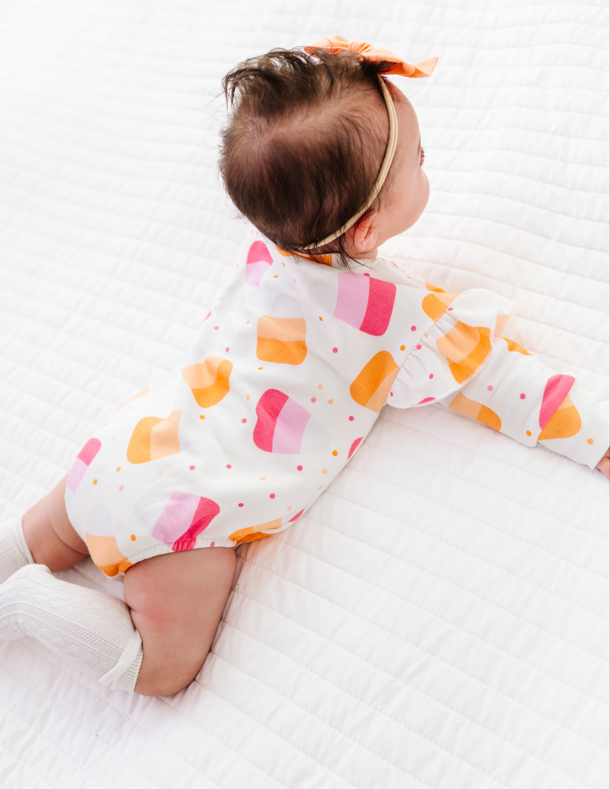 A baby wearing a white long-sleeve romper with a colorful pattern of pink, orange, and yellow hearts, lying on a white quilted blanket.
