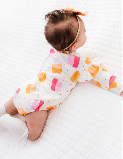 A baby wearing a white long-sleeve romper with a colorful pattern of pink, orange, and yellow hearts, lying on a white quilted blanket.