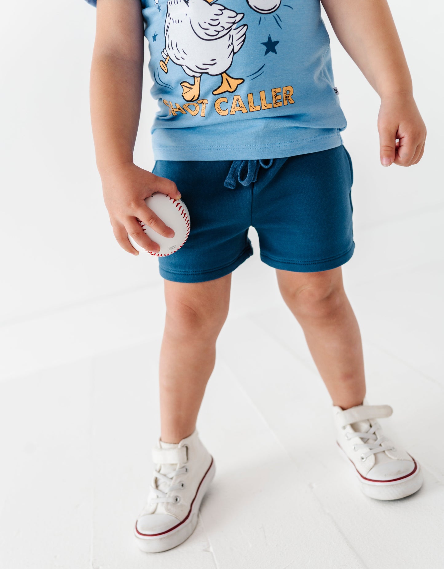 Child wearing a blue shirt with a cartoon design and blue shorts, holding a baseball on a white background