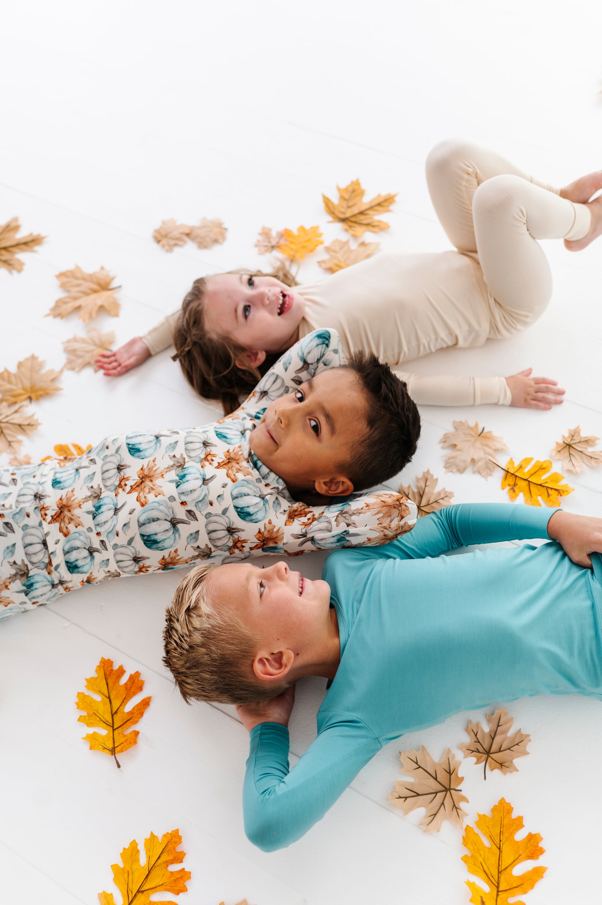 Three children lying on a white surface with autumn leaf decorations wearing bamboo pajamas