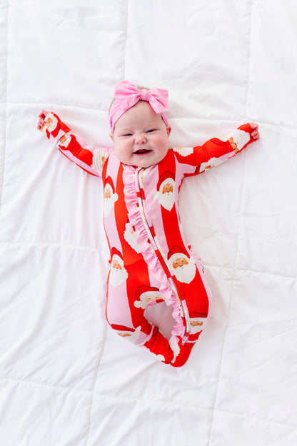 A baby lying on a white quilted blanket wearing a red and pink Santa Claus print footed bamboo sleeper with a pink bow headband.