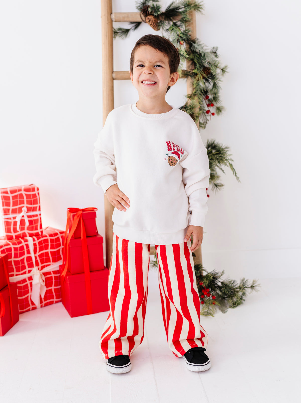 A young boy stands smiling in a holiday-themed room, wearing a white sweatshirt with a small Santa graphic on the chest and red-and-white vertically striped pants. Red wrapped gifts and a decorated Christmas ladder are visible in the background.