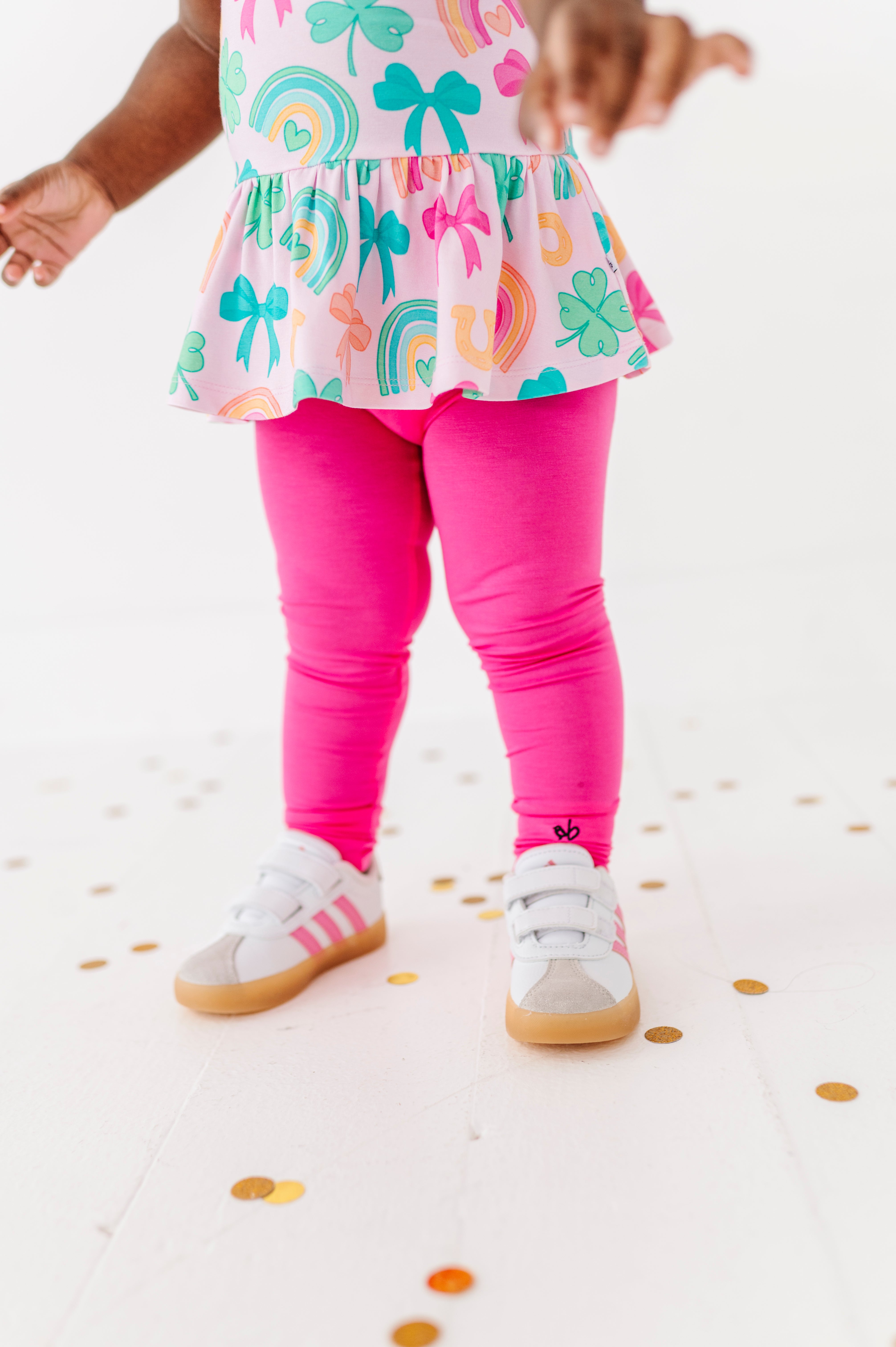 Child wearing a colorful top with rainbow and bow pattern and pink leggings on a white background.