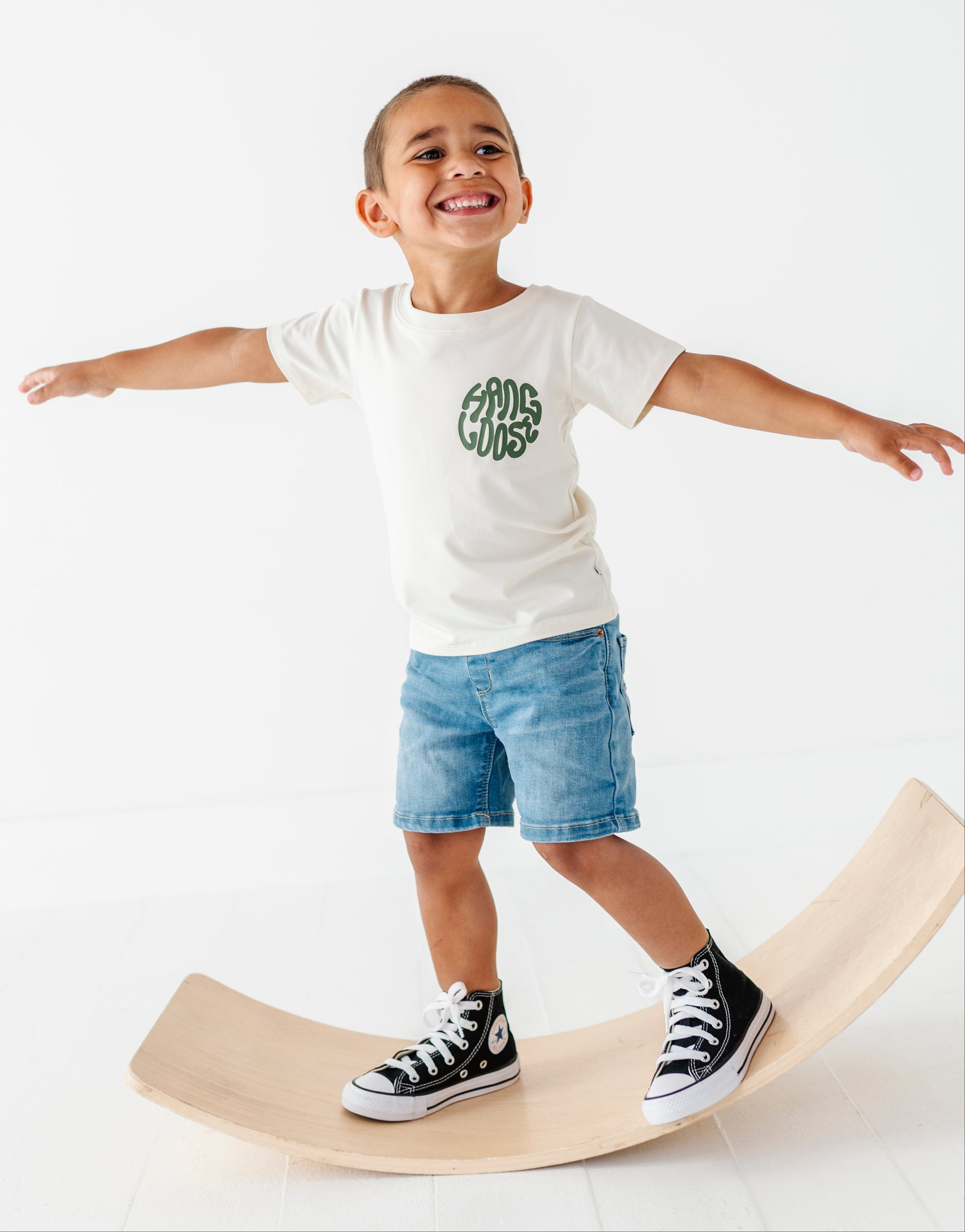 Child standing on a wooden balance board wearing a white t-shirt with green text and blue shorts.