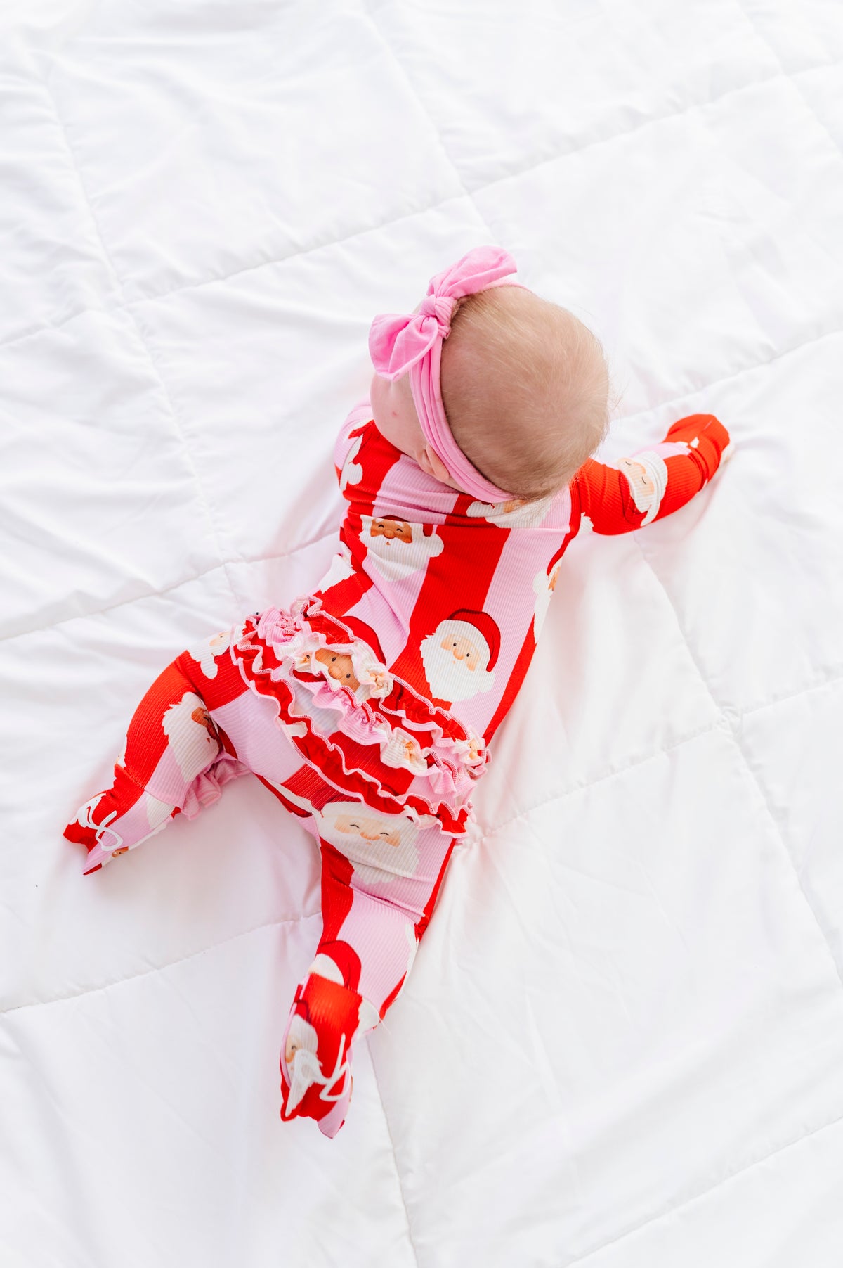 A baby lying on a white quilted blanket, wearing a festive red and white Santa Claus print footed sleeper with a ruffled hem, a pink bow headband, and red socks.