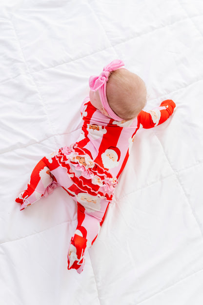 A baby lying on a white quilted blanket, wearing a festive red and white Santa Claus print footed sleeper with a ruffled hem, a pink bow headband, and red socks.